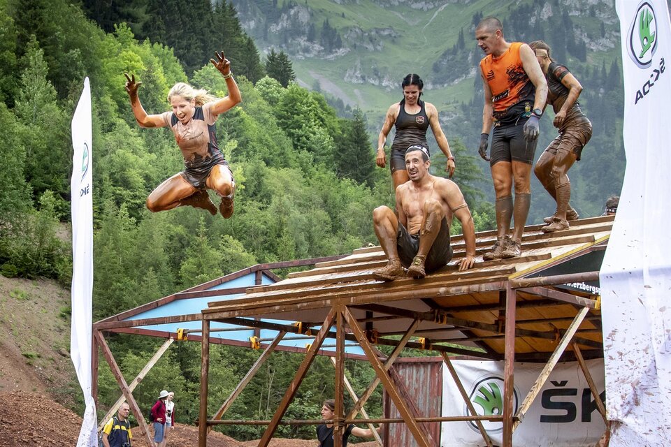 Eine Gruppe von Menschen nimmt an einem Hindernisrennen teil und springt von einer Plattform. Die Teilnehmer sind dreckig und haben viel Spaß in einer natürlichen Umgebung. | © August Zoebl