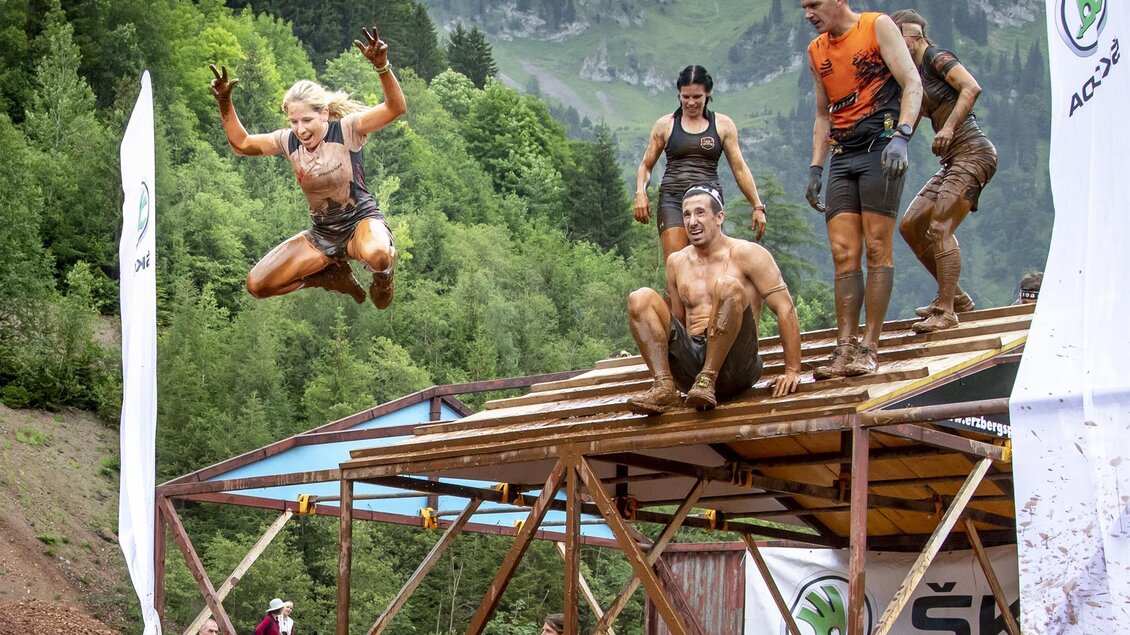Eine Gruppe von Menschen nimmt an einem Hindernisrennen teil und springt von einer Plattform. Die Teilnehmer sind dreckig und haben viel Spaß in einer natürlichen Umgebung. | © August Zoebl
