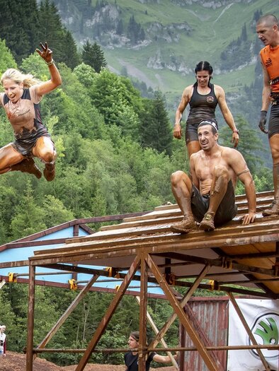 A group of people is participating in an obstacle race and jumping off a platform. The participants are dirty and having a lot of fun in a natural environment. | © August Zoebl