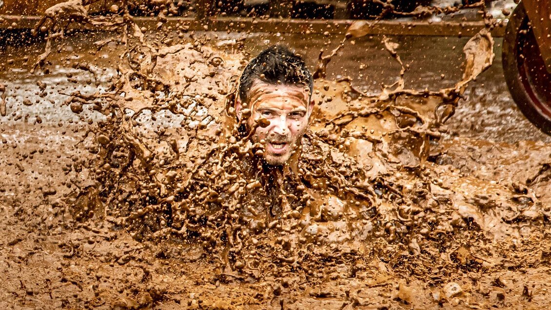 Ein Mann taucht in eine Spritzwasserschlacht aus Schlamm. Um ihn herum fliegen Schlammspritzer in die Luft. | © August Zoebl