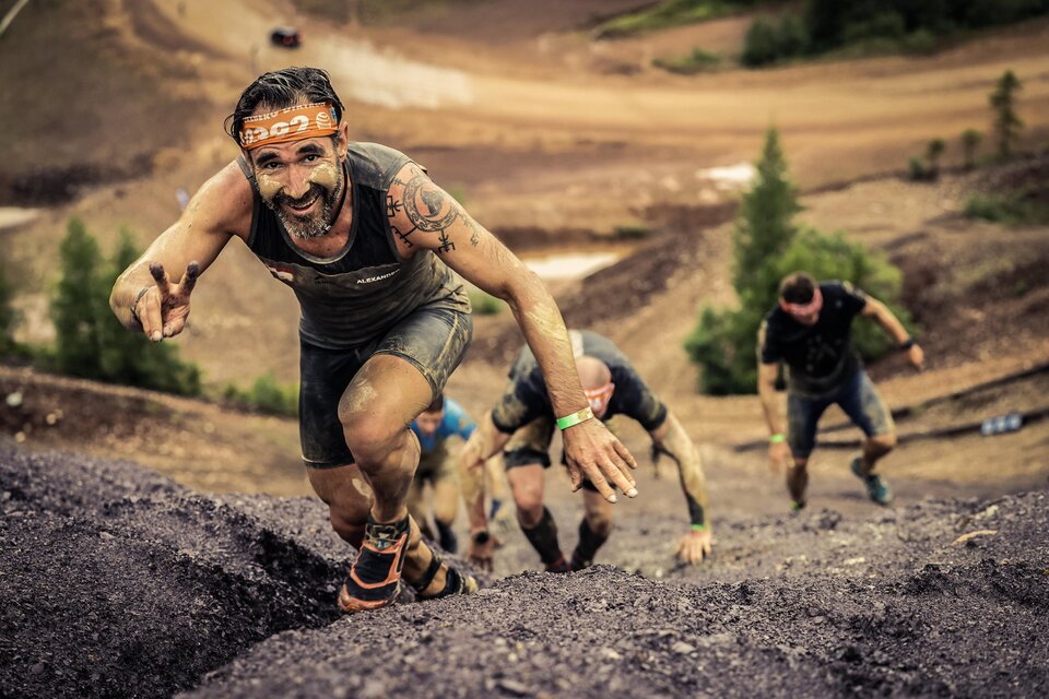 Eine Gruppe von Männern klettert an einem steilen Hang. Einer der Männer lächelt und gibt das Peace-Zeichen. | © Sportfotograf Tiqa