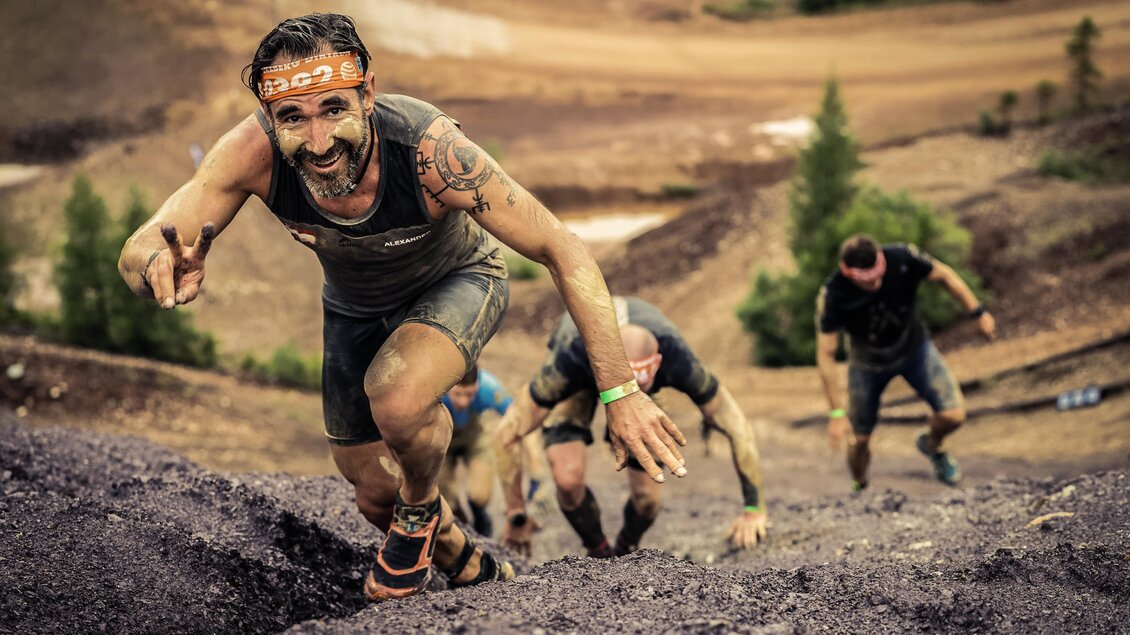 Eine Gruppe von Männern klettert an einem steilen Hang. Einer der Männer lächelt und gibt das Peace-Zeichen. | © Sportfotograf Tiqa
