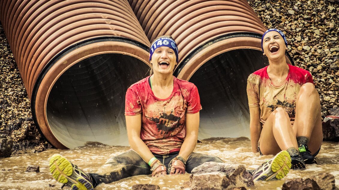 Zwei Frauen sitzen lachend im schlammigen Wasser vor großen Rohren. Beide sind schmutzig und tragen Sportkleidung sowie Kopfband. | © Sportfotograf Tiqa