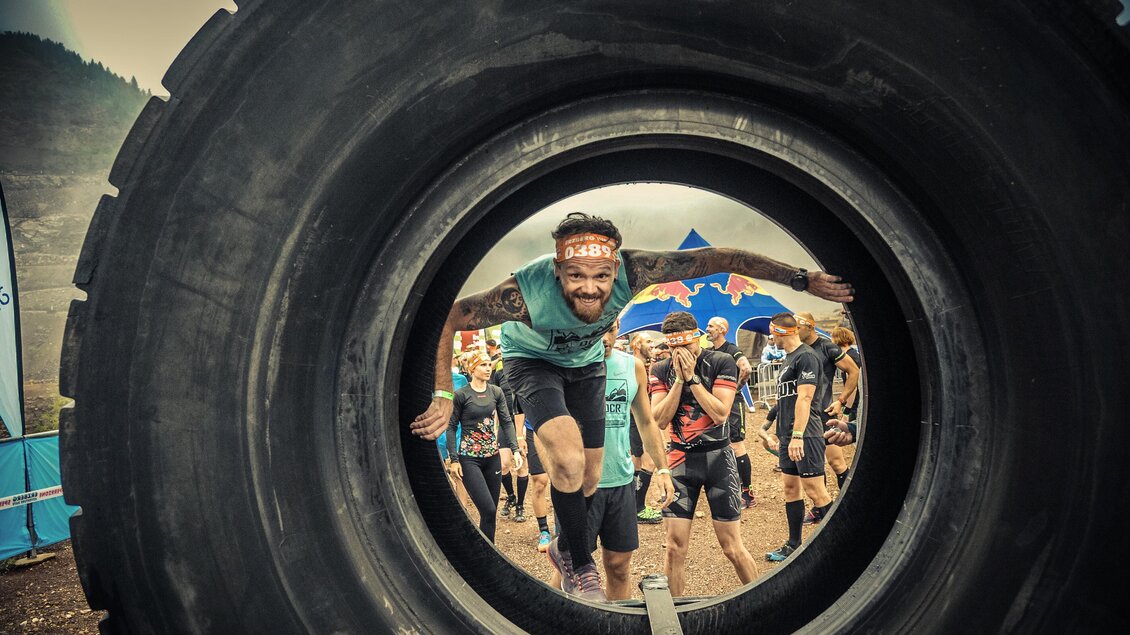 Ein Teilnehmer überwindet ein großes Reifenhindernis während eines Wettkampfs. Im Hintergrund sind weitere Teilnehmer und eine bunte Kulisse zu sehen. | © Sportfotograf Tiqa
