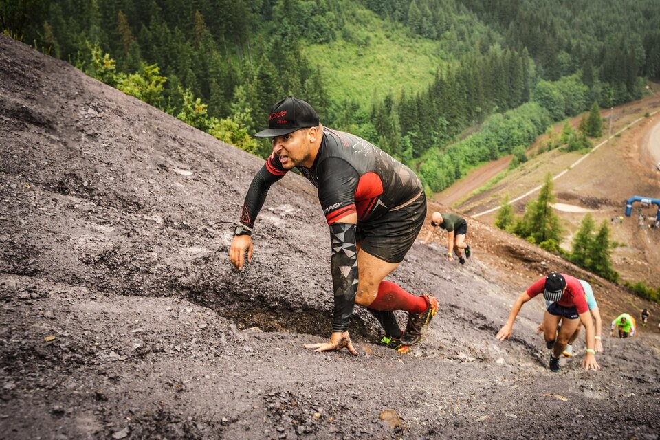 Eine Gruppe von Männern klettert einen steilen, schlammigen Hang hinauf. Die Umgebung ist grünes Gebirge mit Bäumen im Hintergrund. | © Tiqa