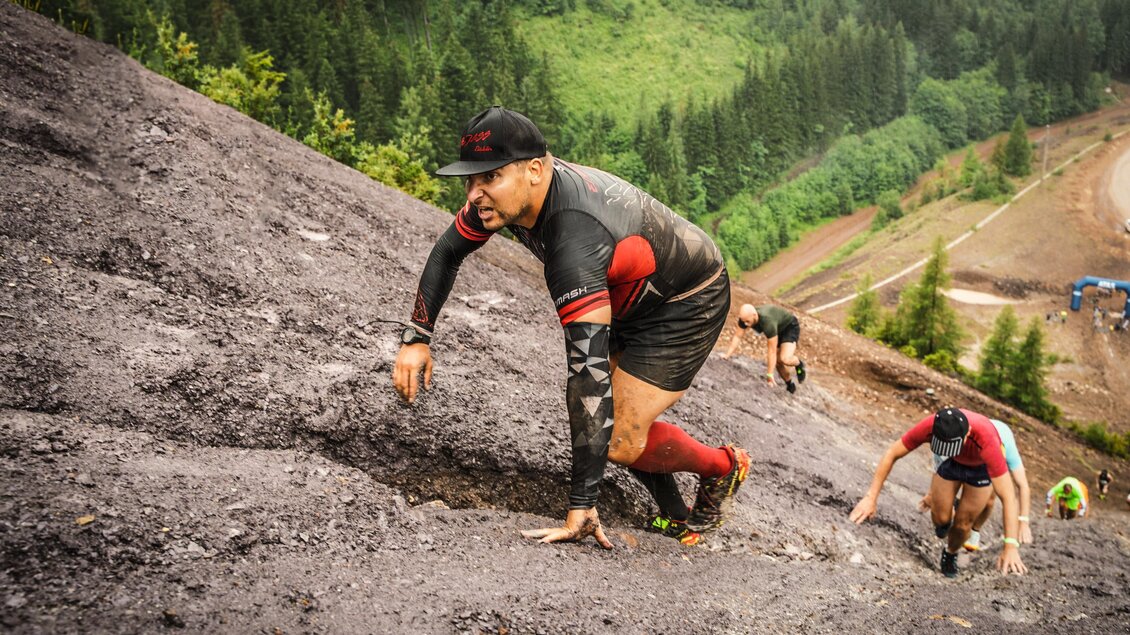 Eine Gruppe von Männern klettert einen steilen, schlammigen Hang hinauf. Die Umgebung ist grünes Gebirge mit Bäumen im Hintergrund. | © Tiqa
