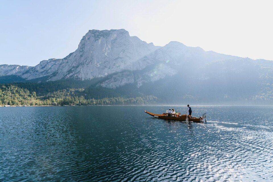 Eine ruhige Seenlandschaft mit einem Fischerboot und hohen Bergen im Hintergrund. Die Sonne strahlt und reflektiert auf dem Wasser. | ©  Seevilla_Karl Steinegger