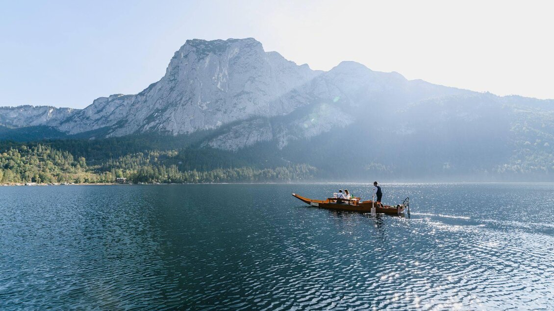 Eine ruhige Seenlandschaft mit einem Fischerboot und hohen Bergen im Hintergrund. Die Sonne strahlt und reflektiert auf dem Wasser. | ©  Seevilla_Karl Steinegger
