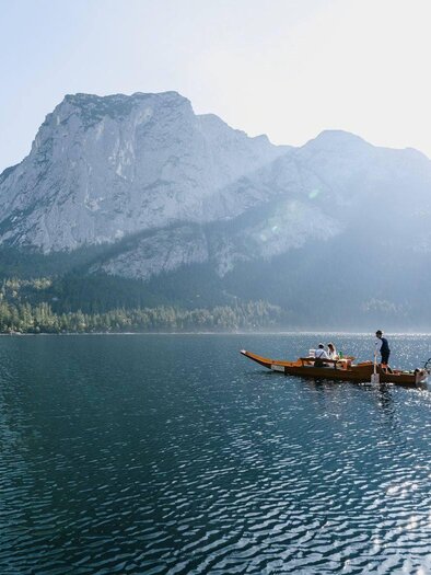 A tranquil lake landscape with a fishing boat and tall mountains in the background. The sun shines and reflects on the water. | ©  Seevilla_Karl Steinegger