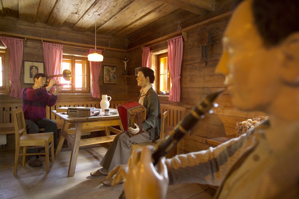 Eine traditionelle Stube mit Holzverkleidung. Zwei Figuren vermitteln eine lebendige Atmosphäre, während eine Person ein Musikinstrument spielt. | © B. Bergmann
