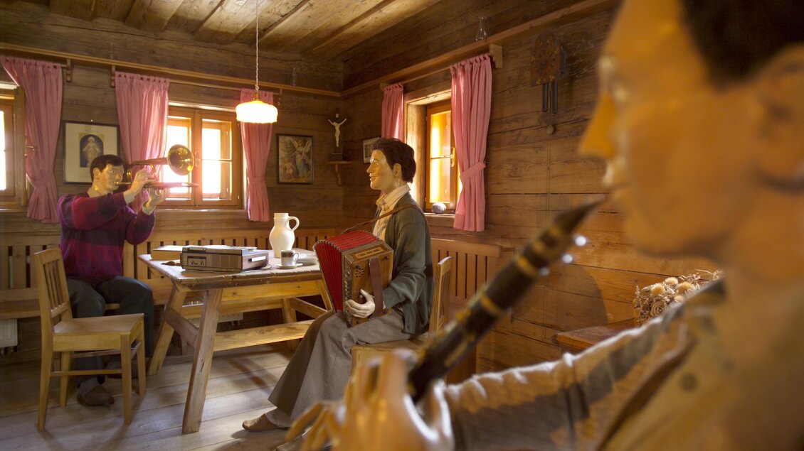 Eine traditionelle Stube mit Holzverkleidung. Zwei Figuren vermitteln eine lebendige Atmosphäre, während eine Person ein Musikinstrument spielt. | © B. Bergmann