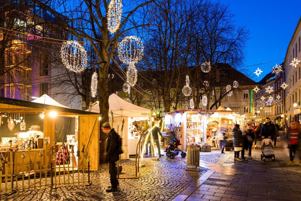 Ein festlicher Weihnachtsmarkt mit bunten Lichtern und Ständen. Besucher schlendern durch die gepflasterte Straße unter einem klaren Abendhimmel. | © Graz Tourismus - Harry Schiffer