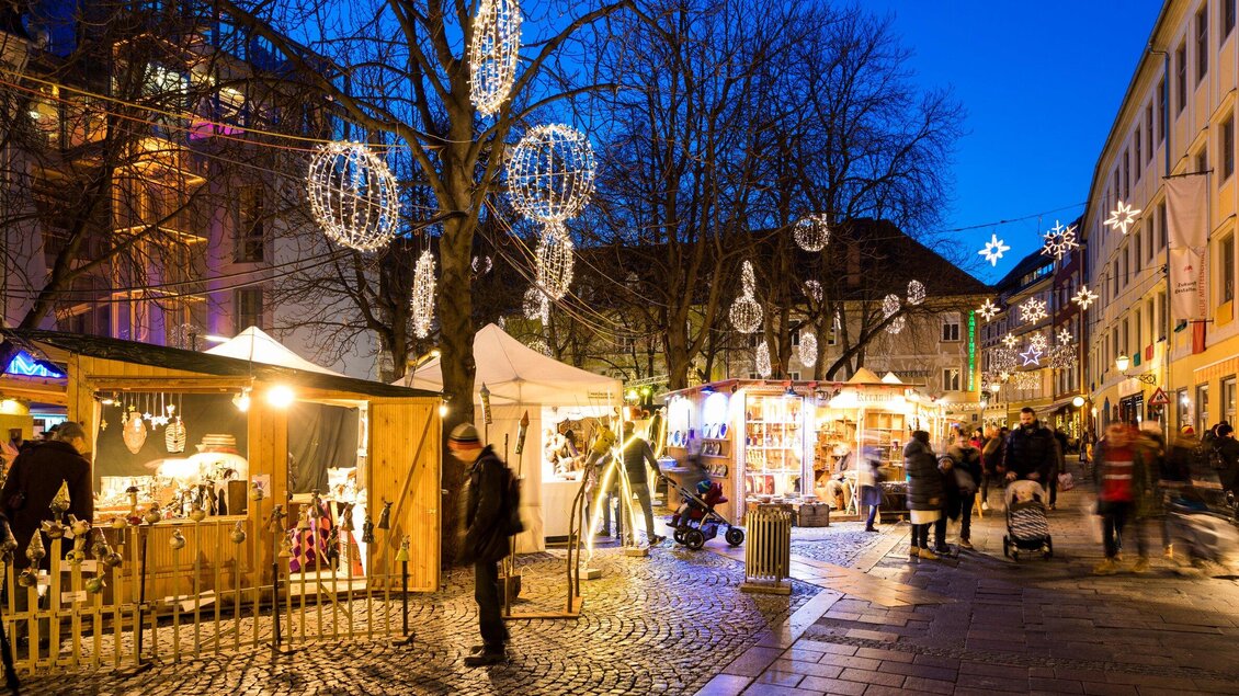 Ein festlicher Weihnachtsmarkt mit bunten Lichtern und Ständen. Besucher schlendern durch die gepflasterte Straße unter einem klaren Abendhimmel. | © Graz Tourismus - Harry Schiffer