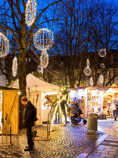 A festive Christmas market with colorful lights and stalls.  
Visitors stroll along the cobblestone street under a clear evening sky. | © Graz Tourismus - Harry Schiffer