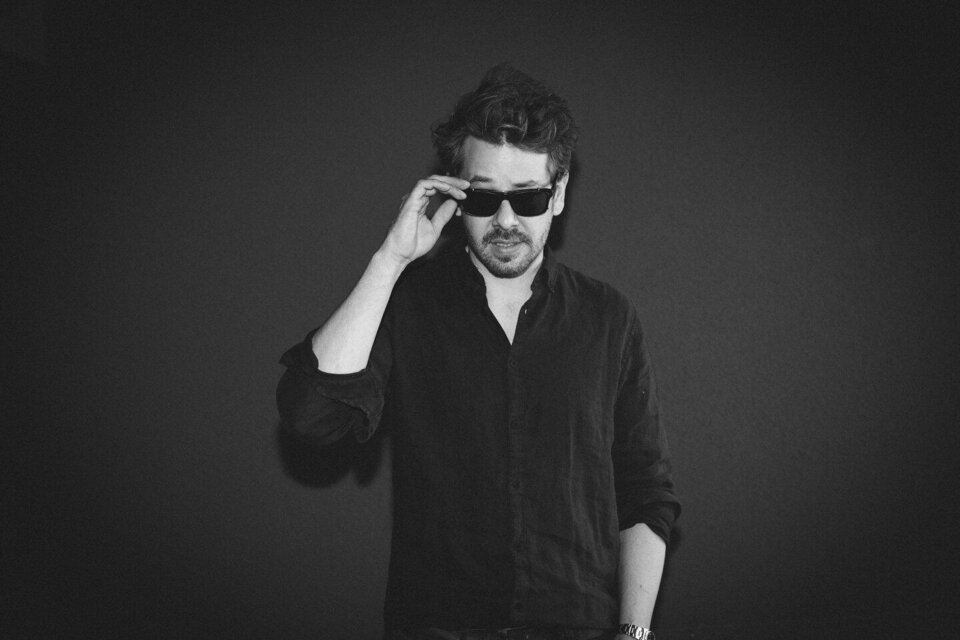 Ein Mann trägt eine schwarze Sonnenbrille und schaut direkt in die Kamera. Er hat lockiges Haar und trägt ein schwarzes Hemd vor einem dunklen Hintergrund. | © LEMO