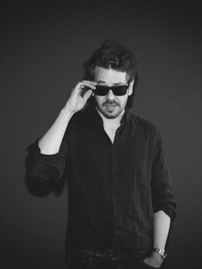 A man is wearing black sunglasses and looking directly at the camera. He has curly hair and is wearing a black shirt against a dark background. | © LEMO