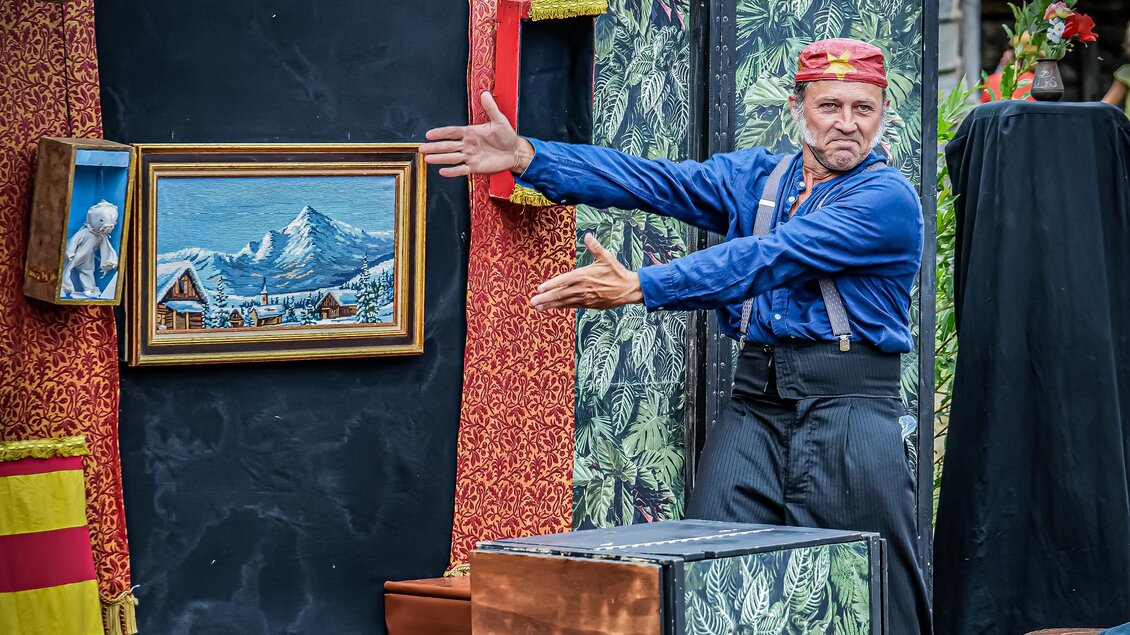 Ein Mann in blauer Kleidung zeigt begeistert auf ein Bild mit Bergen. Im Hintergrund sind bunte Vorhänge und Kisten sichtbar. | © c_Commune_de_Verrieres_en_Anjou
