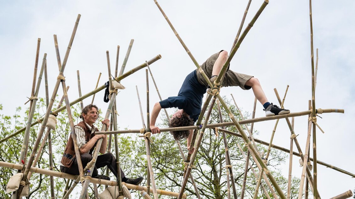 Zwei Personen spielen auf einer Konstruktion aus Holzstäben im Freien. Eine Person balanciert auf den Stangen, während die andere zusieht. | © c_Mark_Robson