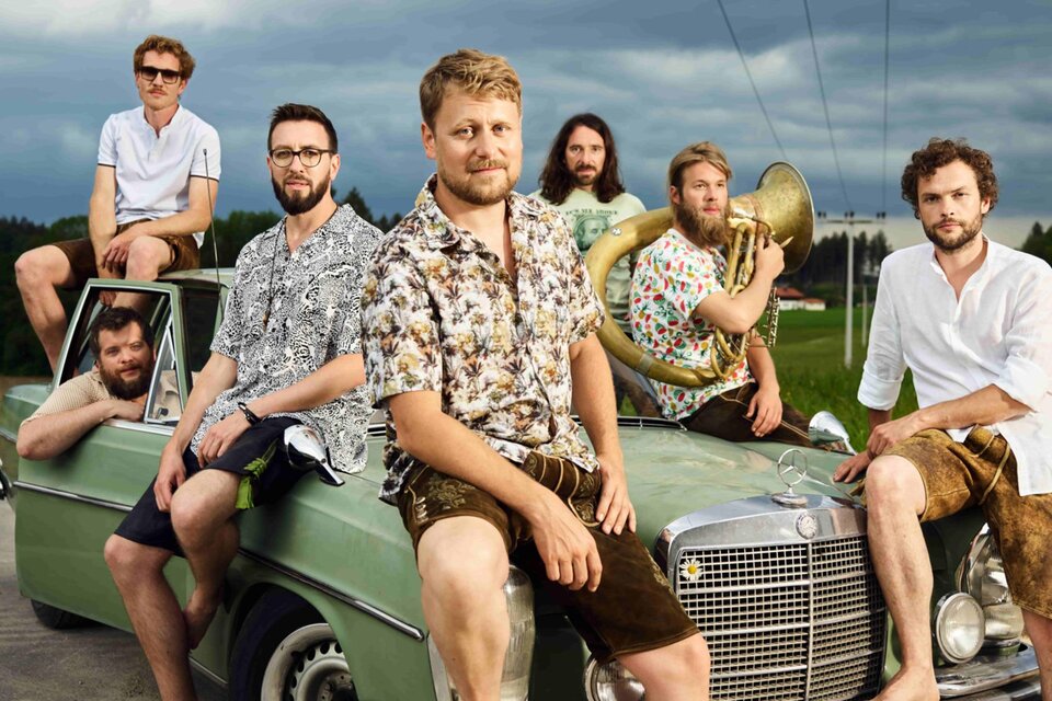 Eine Gruppe von sieben Männern in auffälligen Hemden sitzt auf einem alten Auto. Der Hintergrund zeigt eine ländliche Straße und eine bewölkte Himmel. | © David Königsmann