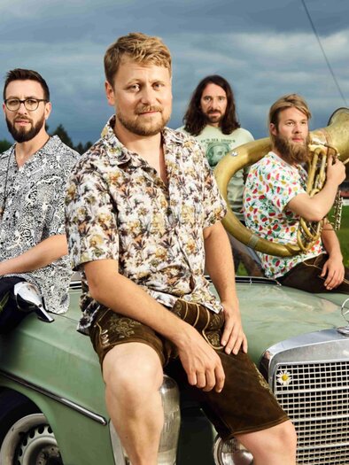 Eine Gruppe von sieben Männern in auffälligen Hemden sitzt auf einem alten Auto. Der Hintergrund zeigt eine ländliche Straße und eine bewölkte Himmel. | © David Königsmann