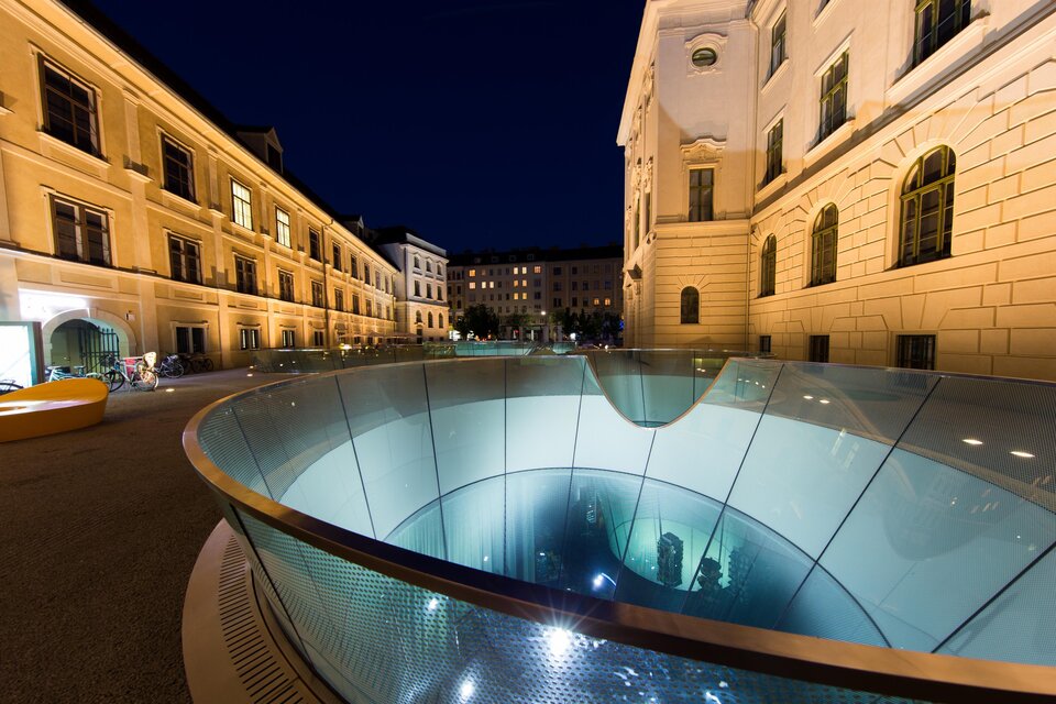 Ein moderner Brunnen mit Glas und Wasser in einer städtischen Umgebung bei Nacht. Die umliegenden Gebäude sind beleuchtet und schaffen eine einladende Atmosphäre. | © (c) Graz Tourismus - Harry Schiffer