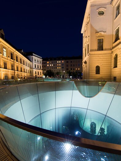 A modern fountain with glass and water in an urban environment at night. The surrounding buildings are illuminated, creating a welcoming atmosphere. | © (c) Graz Tourismus - Harry Schiffer