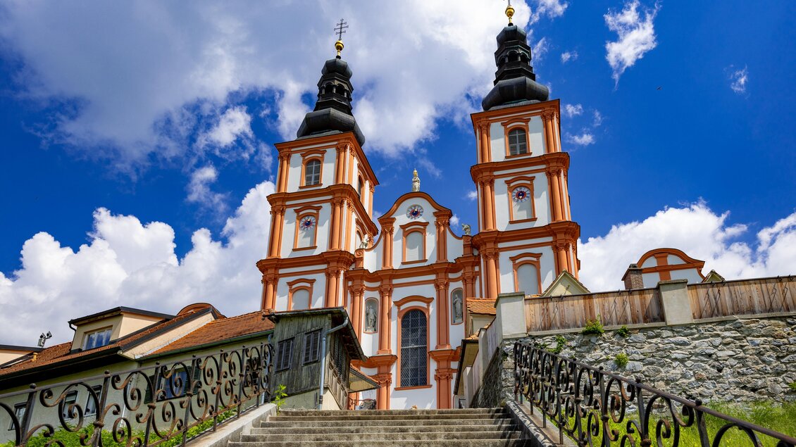 Eine beeindruckende Kirche mit zwei Türmen und bunten Verzierungen. Der Himmel ist blau mit einigen Wolken und die Treppe führt zum Eingang. | © Graz Tourismus-Harry Schiffer