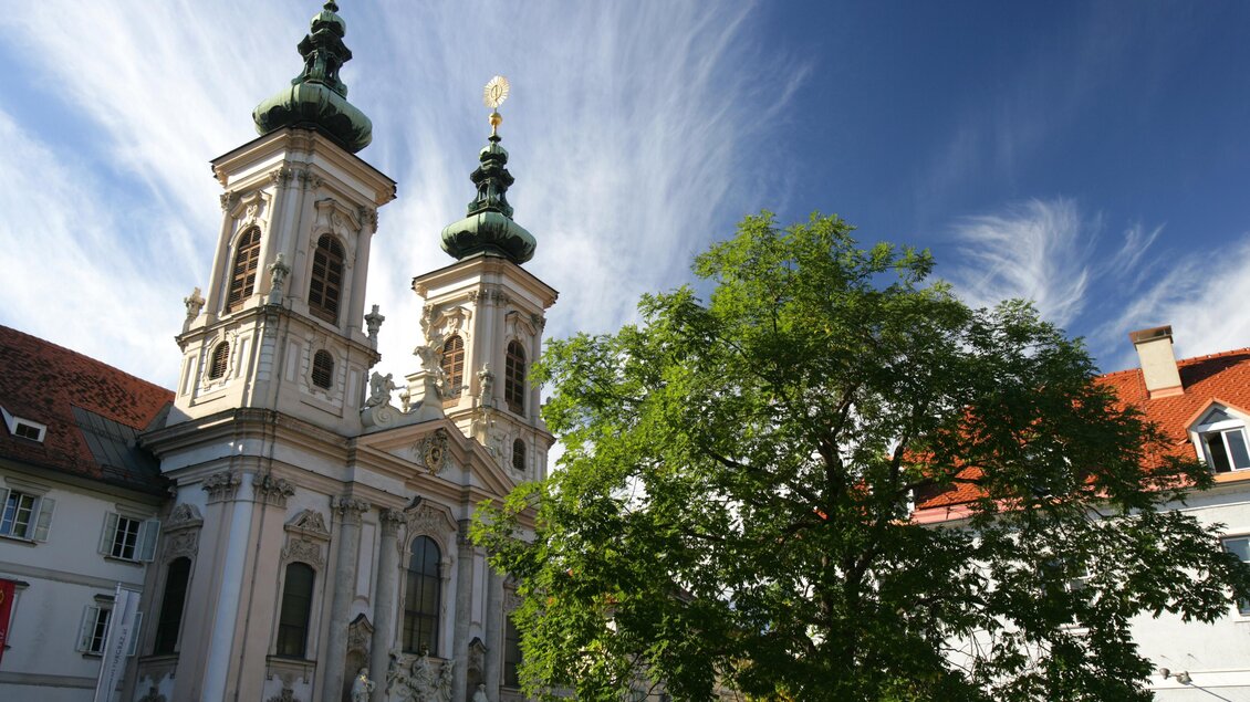 Eine beeindruckende Kirche mit zwei Türmen und einer schönen Fassade. Im Vordergrund steht ein großer Baum vor einem klaren Himmel. | © Graz Tourismus-Harry Schiffer