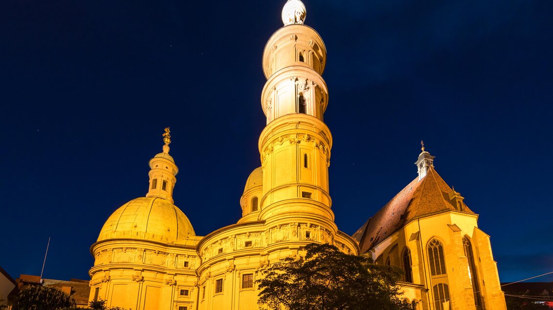 Eine beeindruckende Kirche bei Nacht, beleuchtet in warmem Licht. Der hohe Turm und die Kuppeln sind markante Elemente der Architektur. | © Graz Tourismus-Harry Schiffer