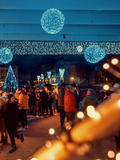 A festive Christmas market with colorful lights and many people. Sparkling decorations hang above the market, creating a cheerful atmosphere. | © MG Lannach - Petru Rimovetz