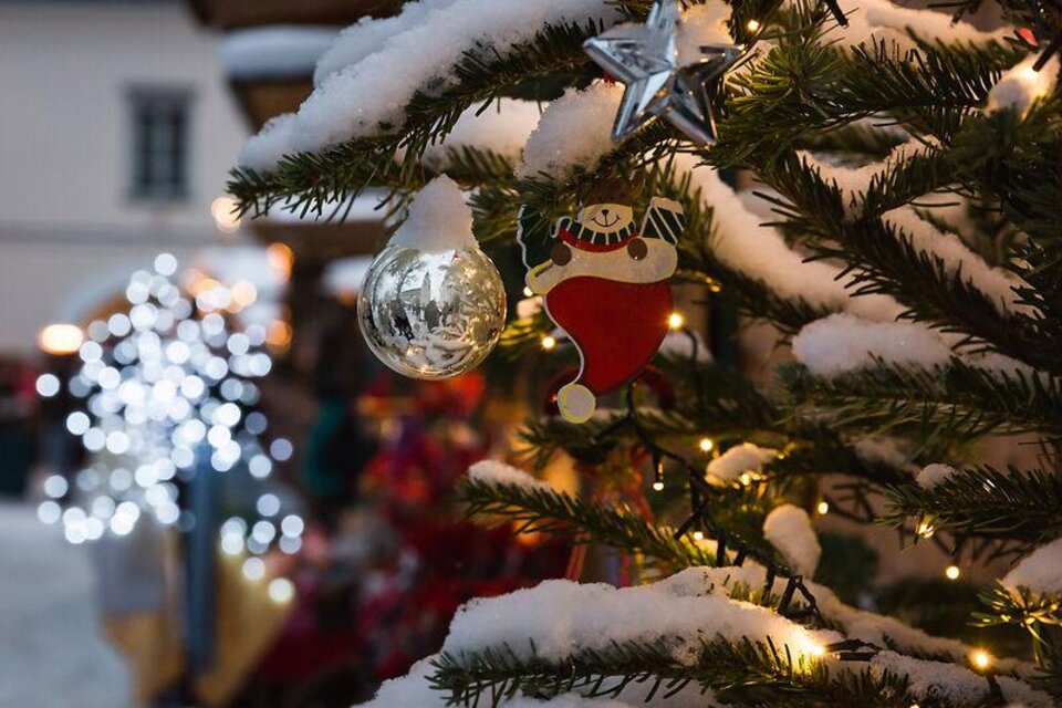 Ein geschmückter Weihnachtsbaum mit Schnee und Lichtern. Im Vordergrund sind eine Christbaumkugel und ein Weihnachtsmann-Anhänger zu sehen.