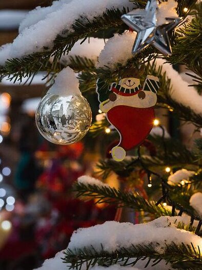 Ein geschmückter Weihnachtsbaum mit Schnee und Lichtern. Im Vordergrund sind eine Christbaumkugel und ein Weihnachtsmann-Anhänger zu sehen.