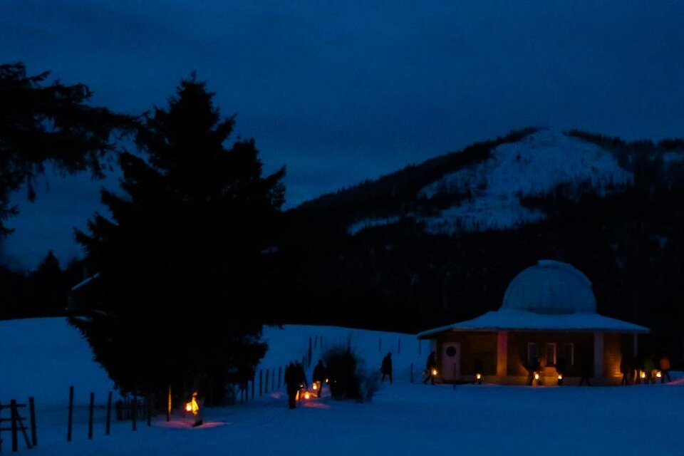 Eine ruhige Winterlandschaft bei Nacht mit einem beleuchteten Gebäude und schneebedeckten Bergen im Hintergrund. Fackeln säumen den Weg und schaffen eine gemütliche Atmosphäre. | © Anna Scherfler