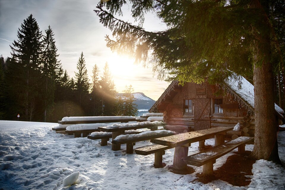 Eine gemütliche Hütte in einer verschneiten Landschaft. Im Vordergrund stehen Holzbänke und der Sonnenuntergang strahlt warmes Licht aus. | © Fred Lindmoser