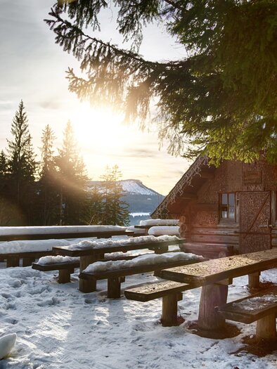 A cozy cabin in a snowy landscape. In the foreground, there are wooden benches, and the sunset radiates warm light. | © Fred Lindmoser