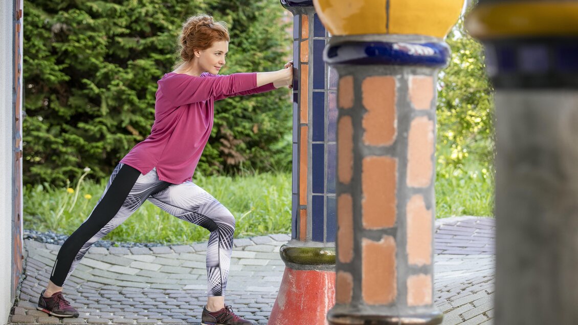 Eine Frau dehnt sich an einer Wand in einem schönen Garten. Sie trägt Sportkleidung und konzentriert sich auf ihre Übung. | © Rogner Bad Blumau(c)Hundertwasser Architketurprojekt