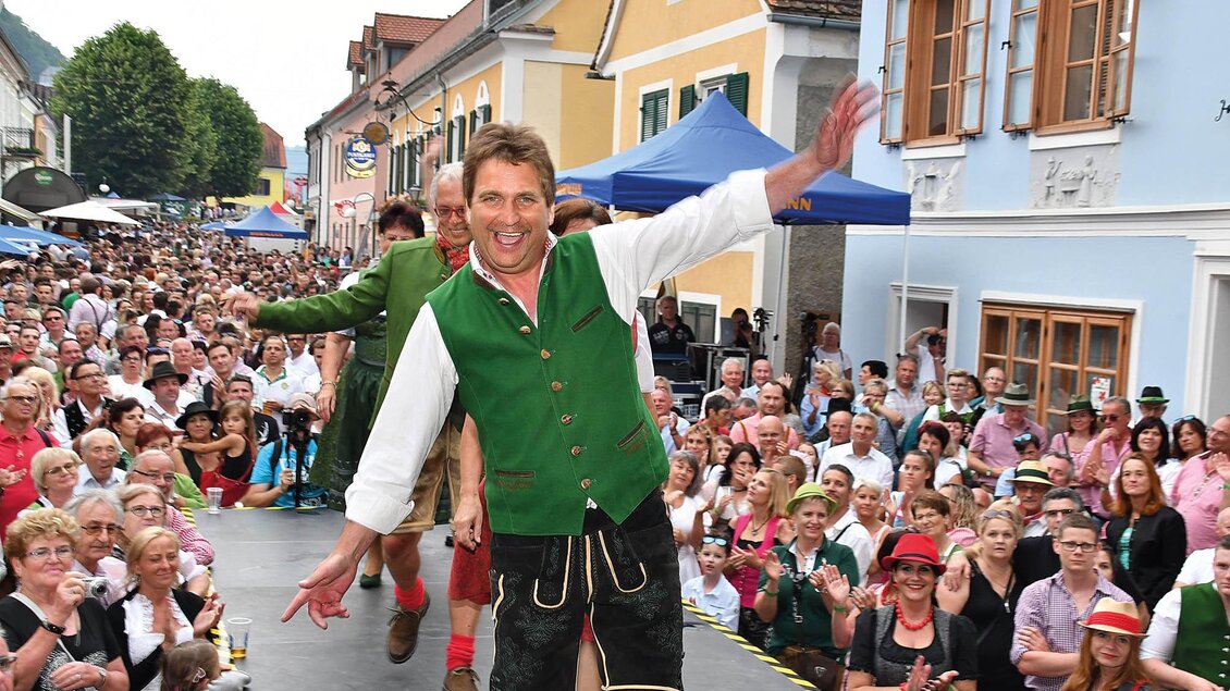 Eine lebhafte Festveranstaltung mit vielen Zuschauern auf der Straße. Ein Mann in traditioneller Tracht tanzt und zieht die Aufmerksamkeit auf sich. | © Hiden-Wagner