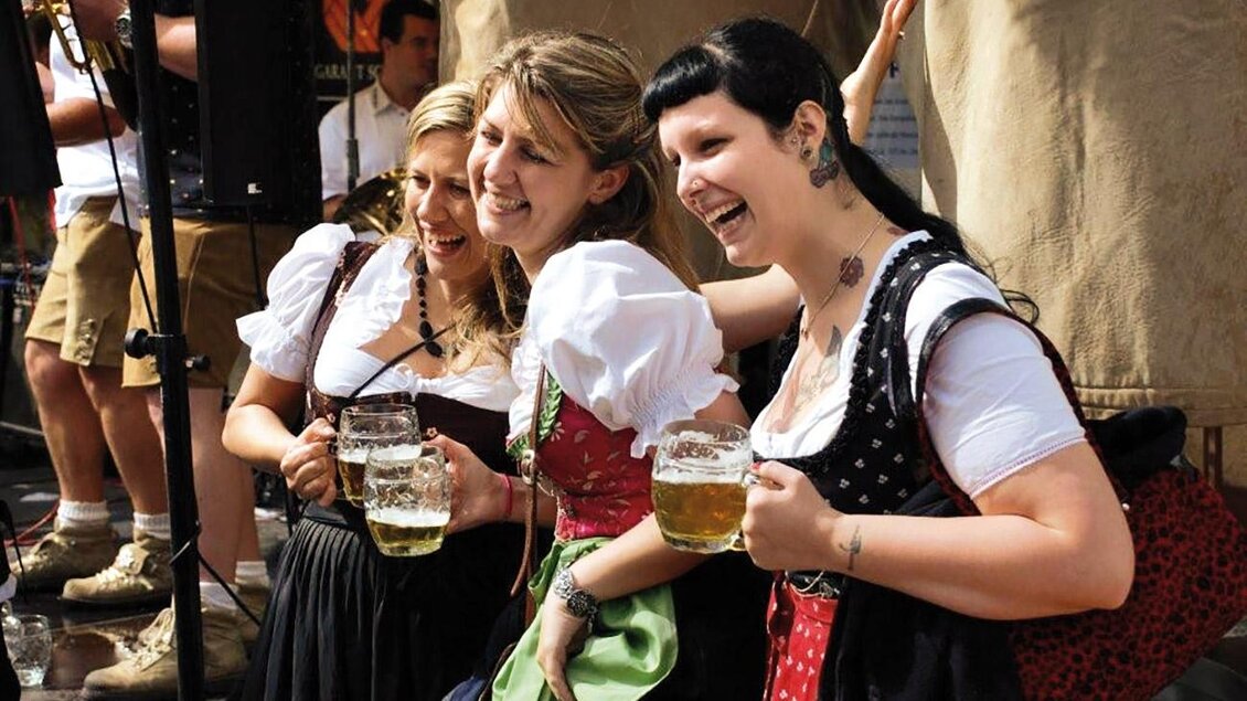 Drei Frauen in traditionellen Trachten feiern fröhlich und halten Bierkrüge in den Händen. Der Hintergrund zeigt eine Feststimmung und eine Menschenmenge. | © Hiden
