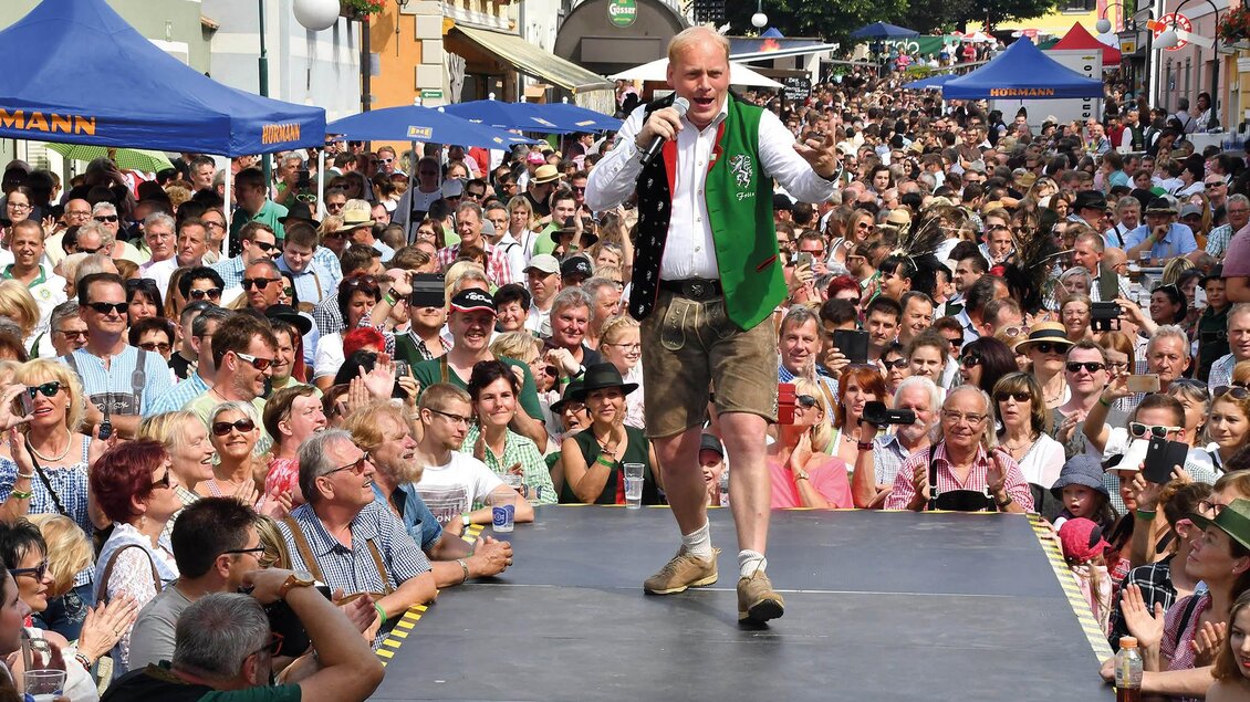 Ein lebhaftes Fest mit einer großen Menschenmenge, die einem Sänger auf einer Bühne zuschaut. Die Veranstaltung findet an einem sonnigen Tag in einer Innenstadt statt. | © Hiden-Wagner