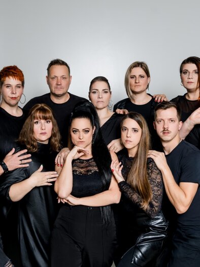 A group of people is assembled in elegant black clothing. They are posing in front of a neutral background, expressive and diverse in their presence. | © Michaela Lorber