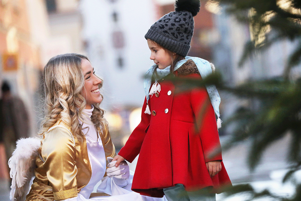 Ein lächelndes Mädchen in einem roten Mantel spricht mit einer Frau in einem goldenen Kostüm. Die Szene spielt sich in einer festlichen Umgebung ab. | ©  Katharina Wassler