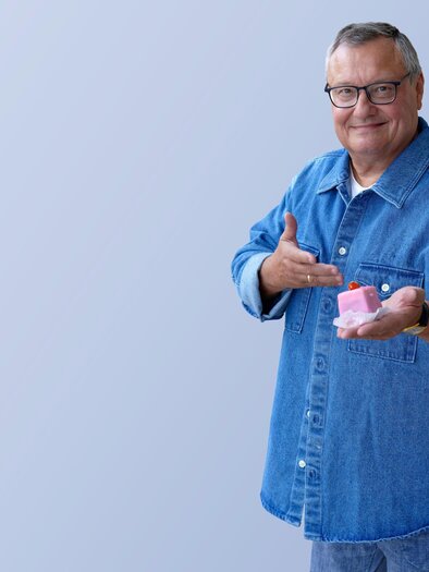 An older man in a blue denim shirt proudly shows a product in his hand. He is smiling and standing in front of a bright background. | © Gary Milano