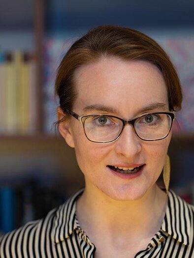 A woman with glasses stands in front of a bookshelf. She is wearing a striped shirt and looking directly at the camera. | ©  Gerald Schutting
