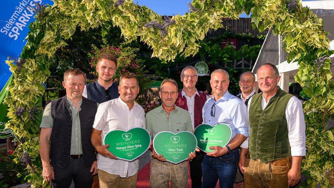 Eine Gruppe von acht Männern steht unter einem herzförmigen Blumenbogen. Sie halten grüne Schilder mit Herzen und lächeln in die Kamera. | © ©pixelmaker