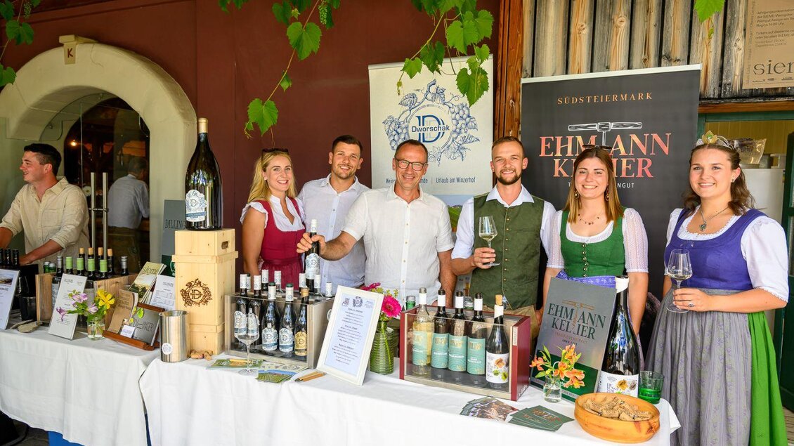 Eine Gruppe von Menschen steht an einem Stand, der verschiedene Weinflaschen und Produkte anbietet. Die Personen tragen traditionelle Kleidung und lächeln freundlich in die Kamera. | © ©pixelmaker