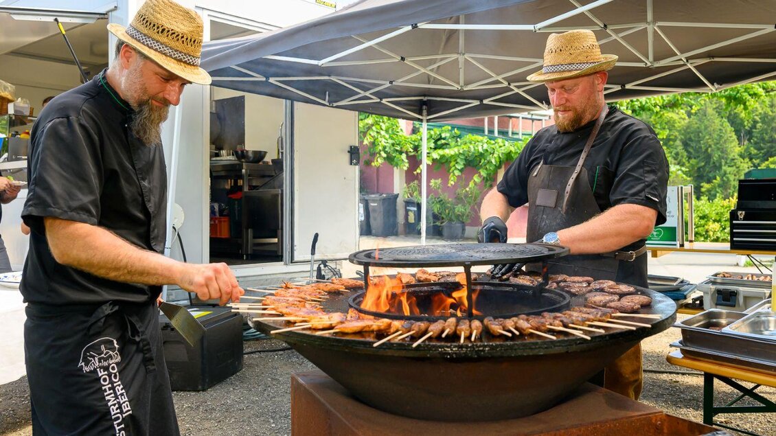 Zwei Köche bereiten Grillgerichte auf einem runden Grill vor. Man sieht eine Vielzahl von Fleischspießen und eine große Flamme. | © ©pixelmaker