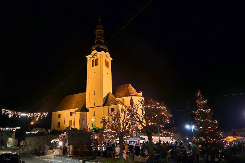 Eine festlich beleuchtete Kirche bei Nacht mit einem Weihnachtsbaum und geschmückten Ständen. Viele Menschen genießen die festliche Atmosphäre. | © Iris Bloder