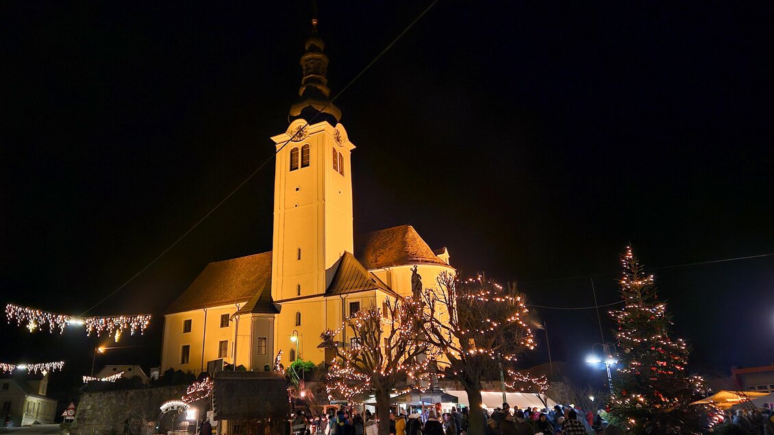 Eine festlich beleuchtete Kirche bei Nacht mit einem Weihnachtsbaum und geschmückten Ständen. Viele Menschen genießen die festliche Atmosphäre. | © Iris Bloder