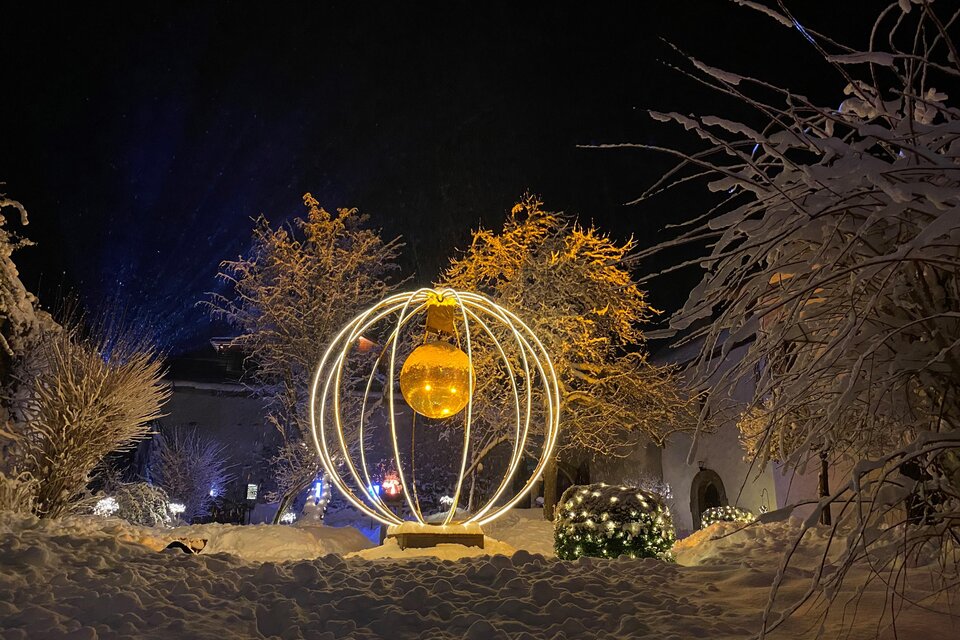 Eine elegante Lichtinstallation in einer verschneiten Winterlandschaft. Die Kugel leuchtet golden und umgeben von schneebedeckten Bäumen und Sträuchern. | © Ulli Koller