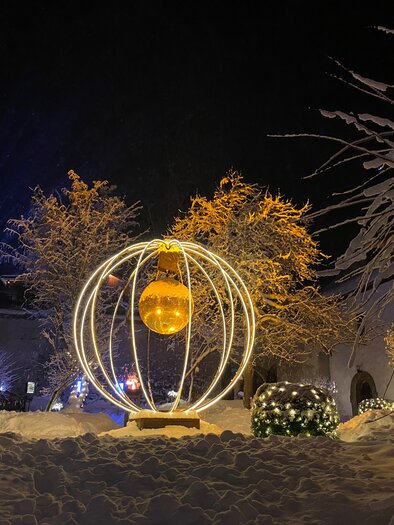 An elegant light installation in a snowy winter landscape. The sphere glows golden and is surrounded by snow-covered trees and shrubs. | © Ulli Koller
